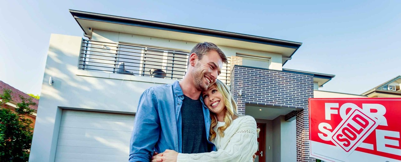 Couple standing in front of a new home. They are standing next to a for sale sign with a sold sticker. They are buying or selling this real estate. They are both wearing casual clothes and embracing. They are smiling and he has a beard. The house is new and contemporary with a brick facade. Copy space